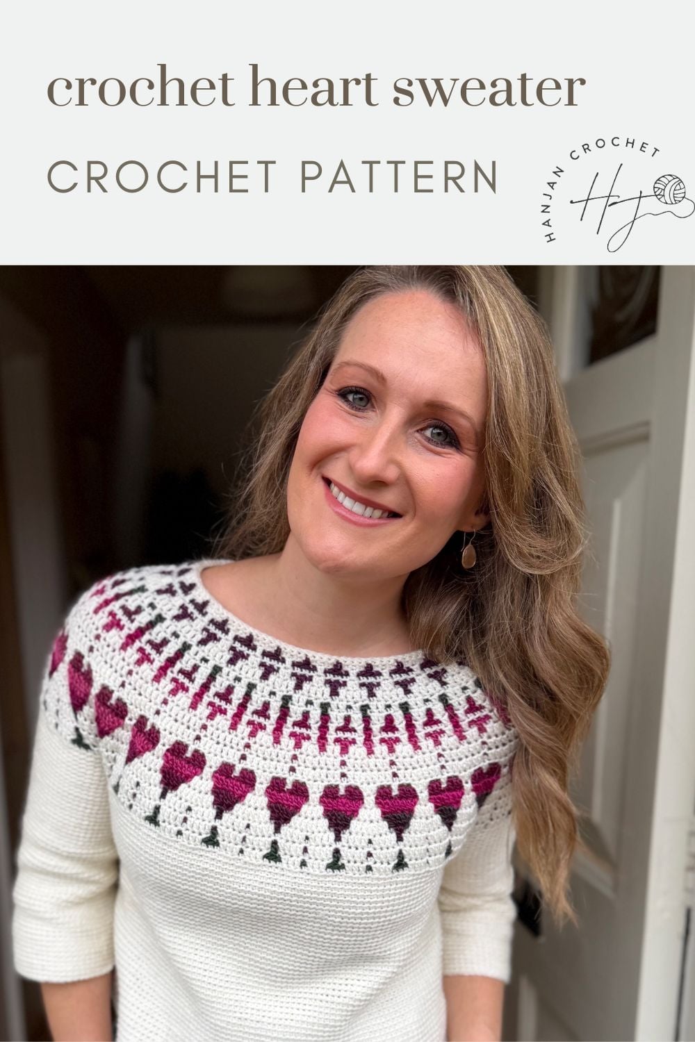 Woman wearing a white crocheted sweater with a pink and green heart pattern near the neckline, standing indoors and smiling at the camera.