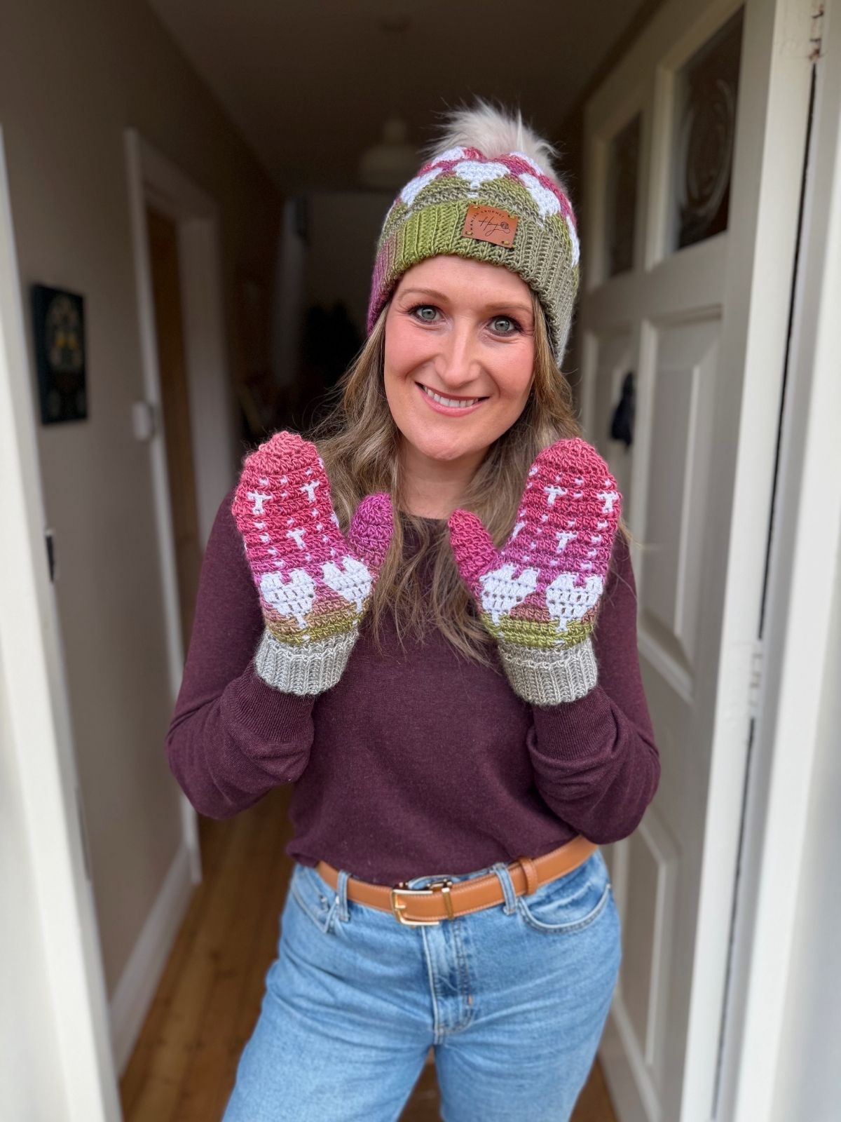 A woman stands indoors, smiling and wearing crochet heart mittens in pink and green tones, paired with a knitted hat, maroon sweater, and blue jeans.