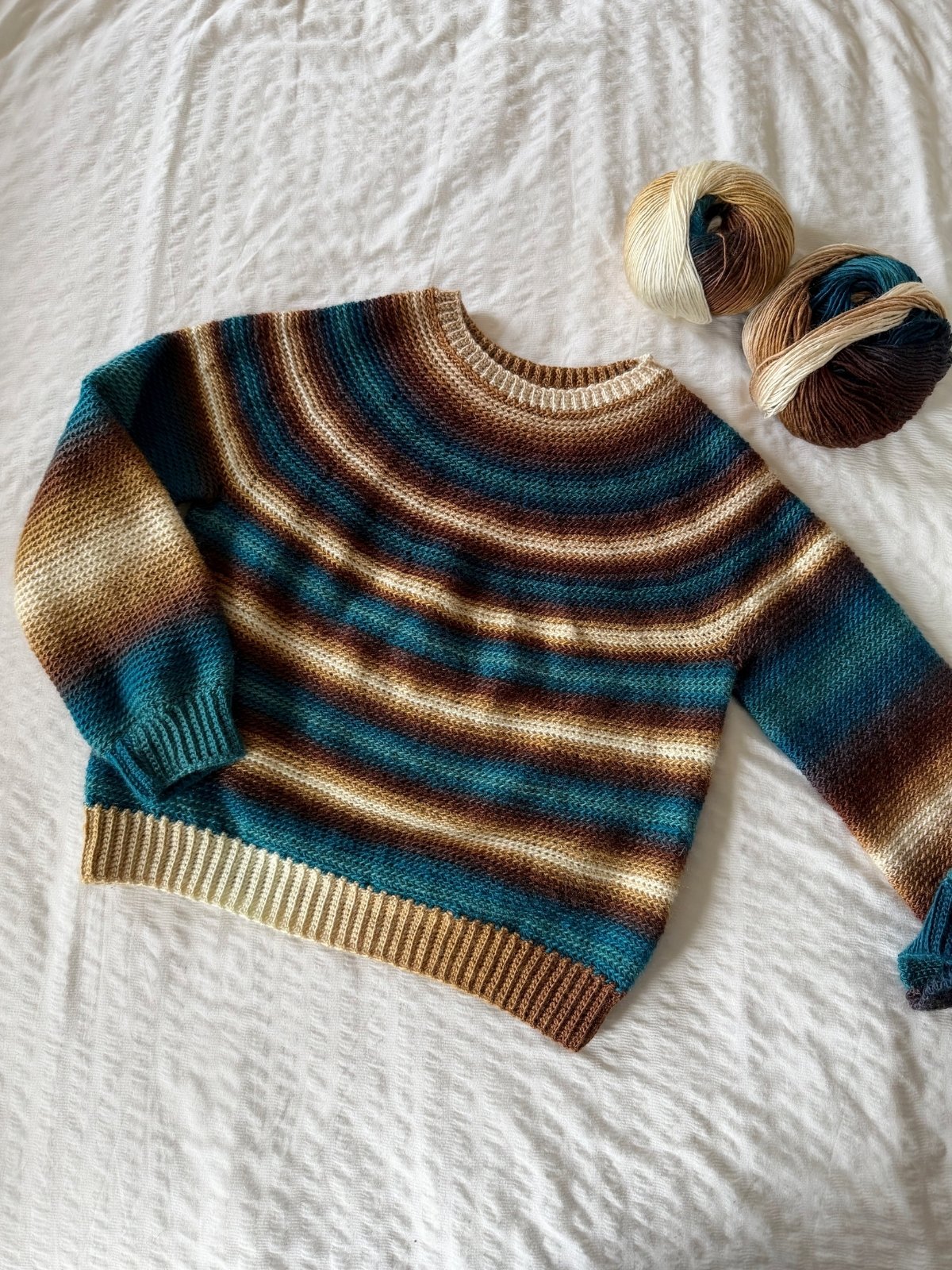 A striped, hand-crocheted sweater in shades of teal, brown, and cream is laid flat on a white textured surface next to two balls of matching yarn.