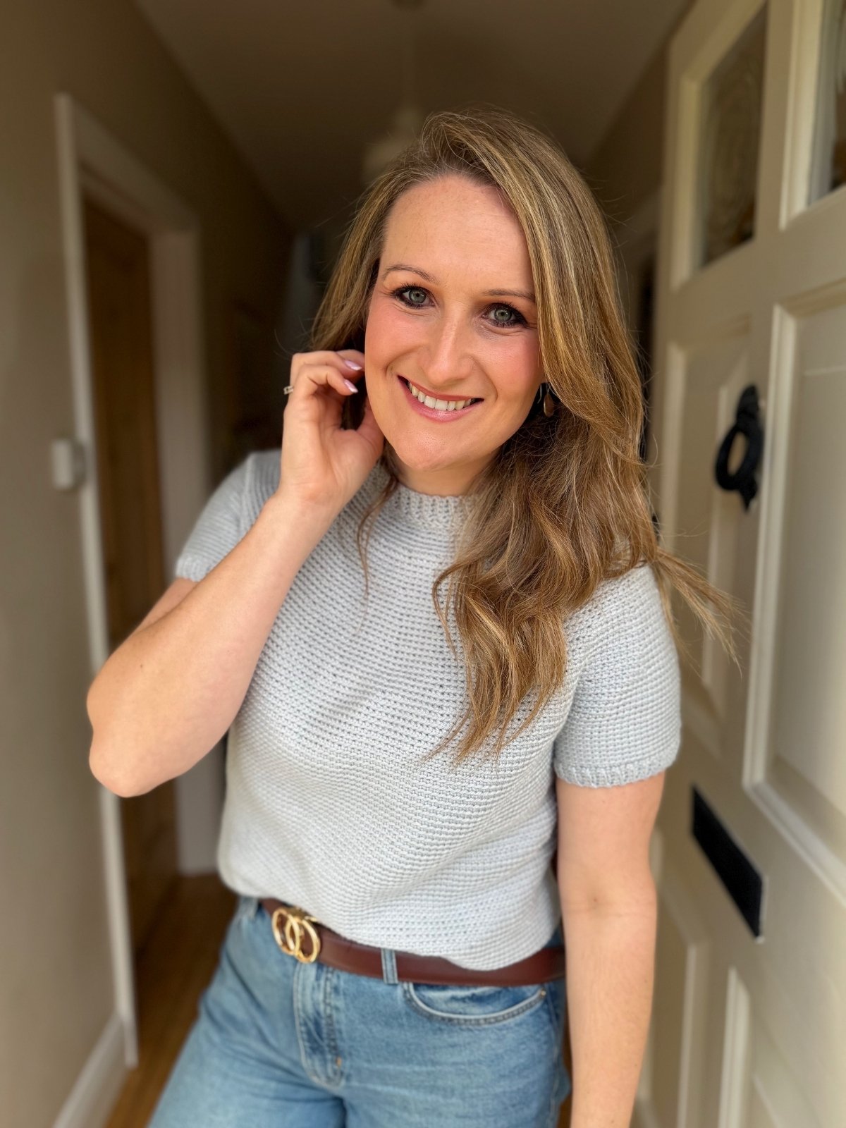 Woman with long blonde hair in a light grey short-sleeve crochet top and blue jeans stands indoors, smiling and touching her hair near a white door.