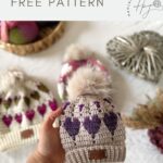 A hand holds a beige crochet hat with purple and lilac heart patterns, topped with a faux fur pom-pom; two similar hats and yarn are in the blurred background, suggesting inspiration from a Free Crochet Heart Hat Pattern.