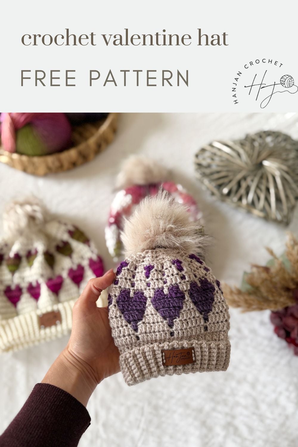 A hand holds a beige crochet hat with purple and lilac heart patterns, topped with a faux fur pom-pom; two similar hats and yarn are in the blurred background, suggesting inspiration from a Free Crochet Heart Hat Pattern.
