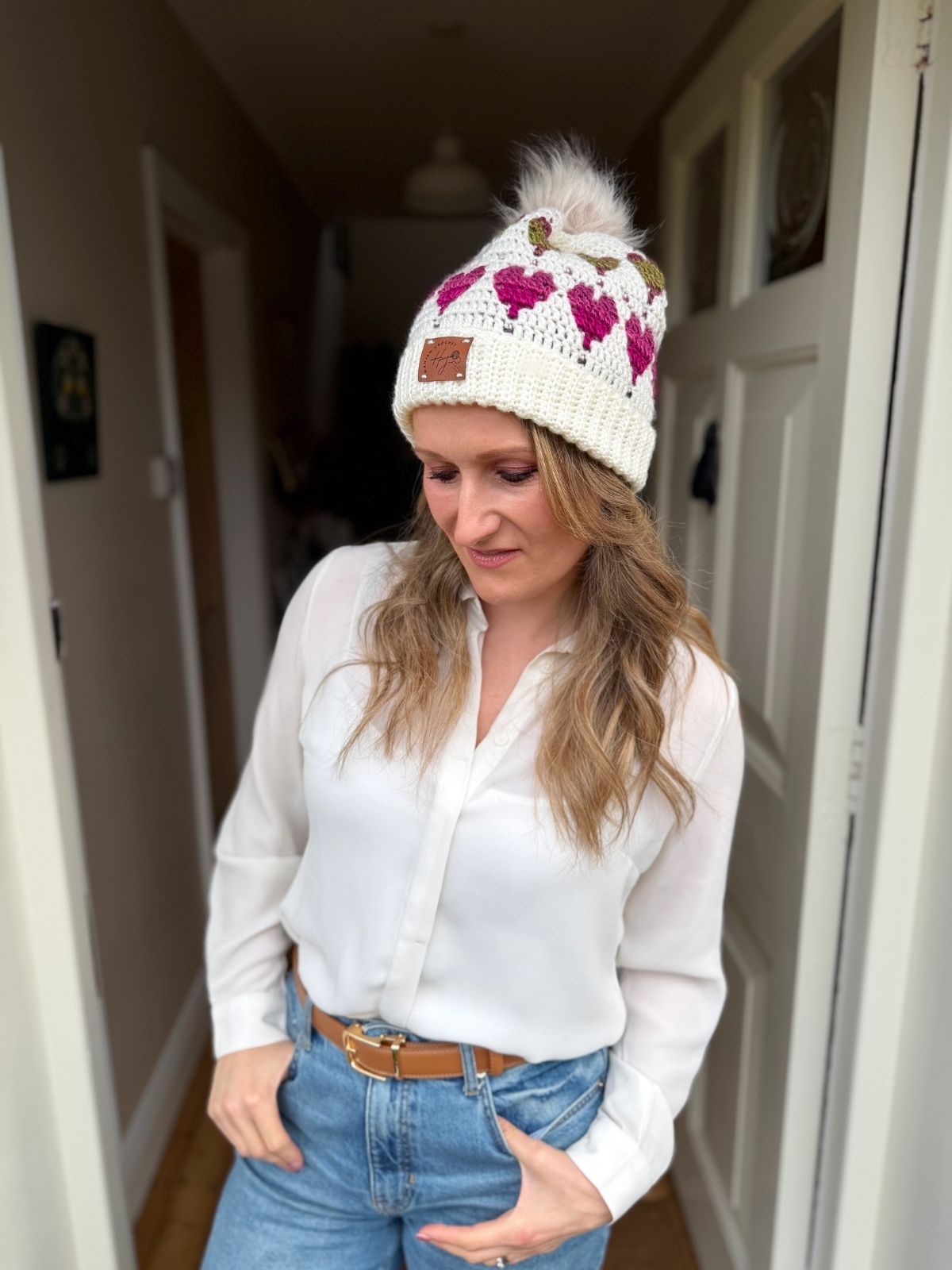 A woman stands in a doorway wearing a white button-up shirt, blue jeans, and a cozy knit hat with pink patterns and a pom-pom, inspired by the Free Crochet Heart Hat Pattern.