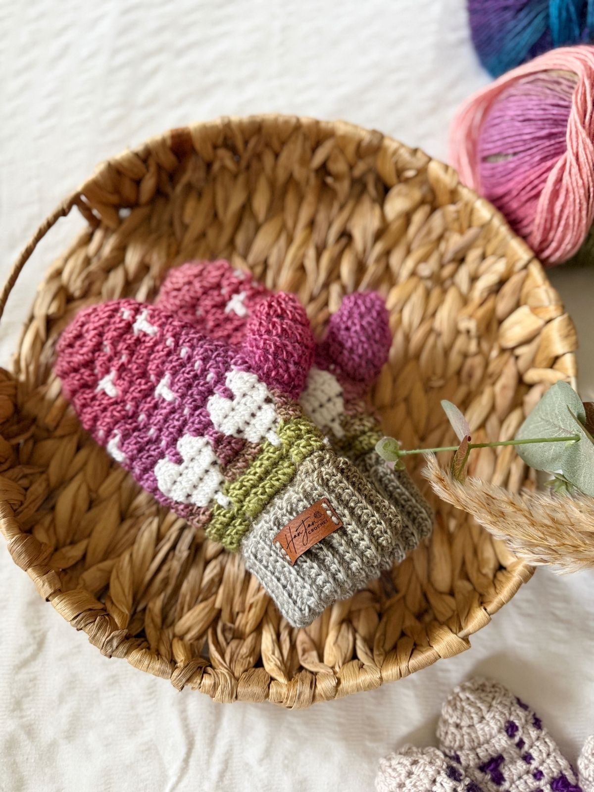 A woven basket holds a pair of pink and white crochet heart mittens with a gray cuff and a tag, next to colorful yarn balls on a white surface.