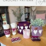 A variety of purple and pink crochet items with white heart patterns are displayed on a table, including baskets, a crochet coffee cozy with heart, and a small pouch.