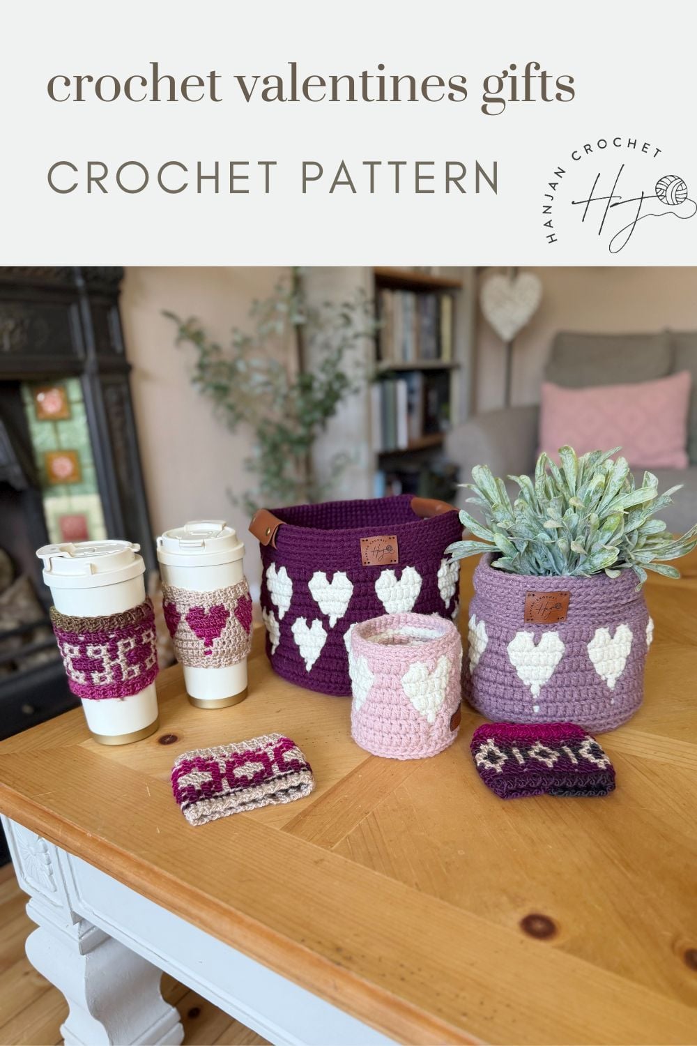 A variety of purple and pink crochet items with white heart patterns are displayed on a table, including baskets, a crochet coffee cozy with heart, and a small pouch.