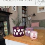 Three crochet baskets with heart patterns in different shades of pink and purple, made using a crochet heart basket pattern, are displayed on a wooden coffee table in a cozy living room.