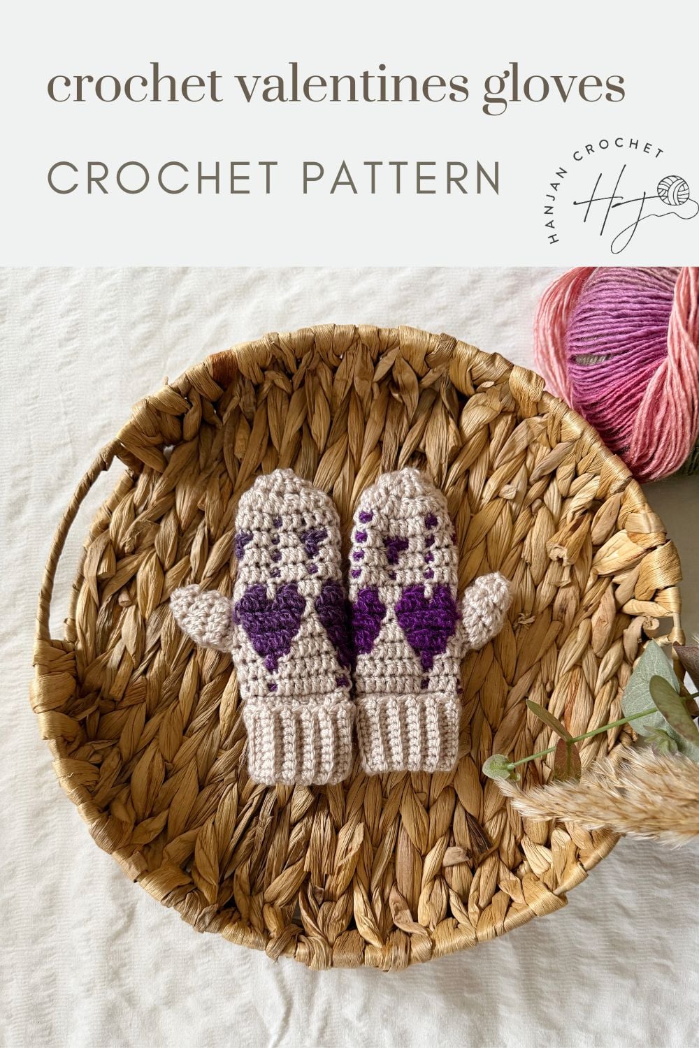 A pair of beige crochet heart mittens with purple heart patterns sits on a woven tray, next to a pink ball of yarn. Text above reads "crochet valentines gloves CROCHET PATTERN".