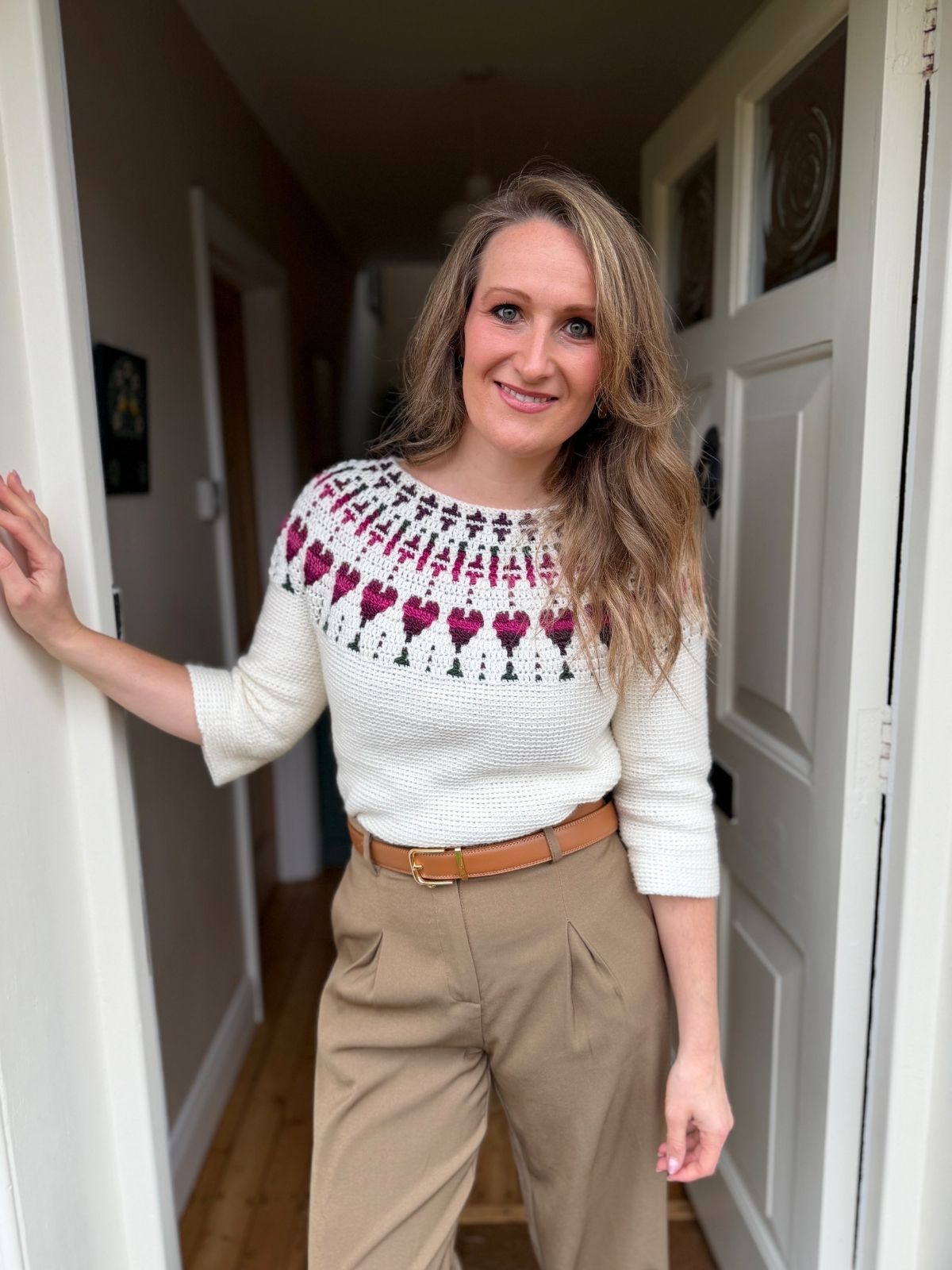 Woman standing in a doorway, wearing a white sweater with a red and purple pattern and tan high-waisted trousers, smiling at the camera.