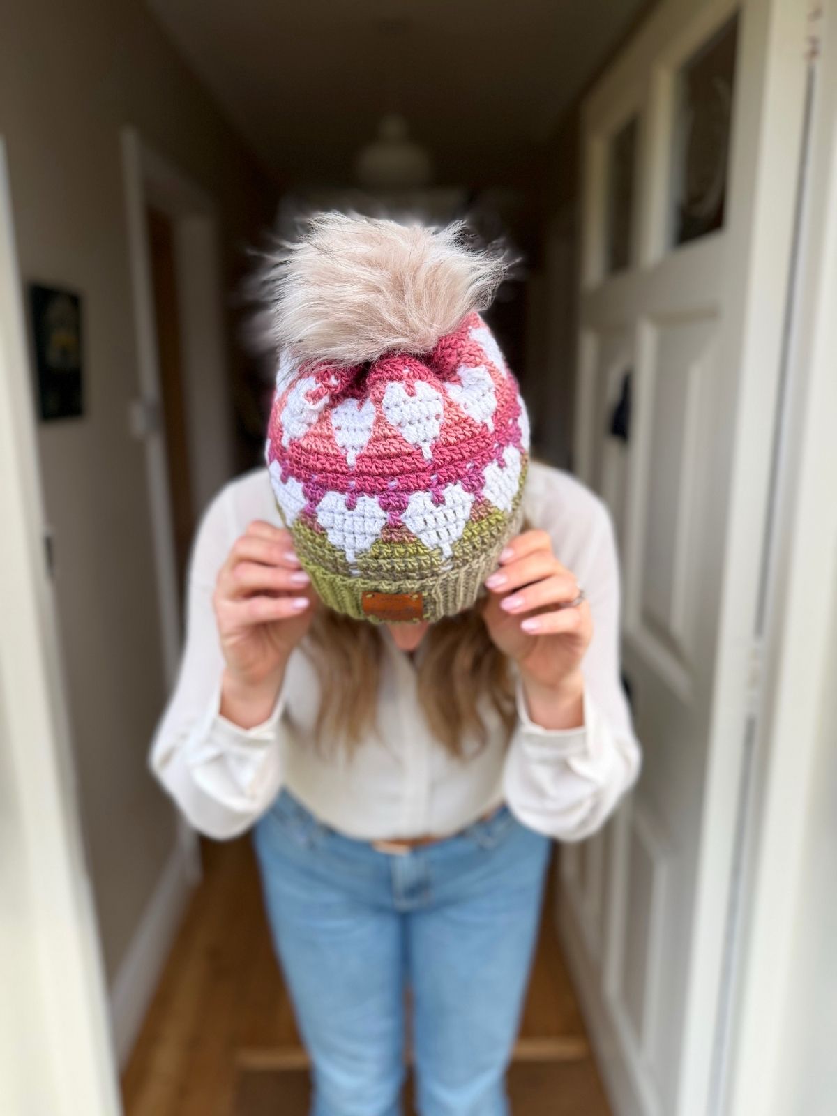 A person wearing a white shirt stands indoors, holding the brim of a knitted beanie with a pom-pom and heart pattern&mdash;perfect inspiration for anyone searching for a Free Crochet Heart Hat Pattern.