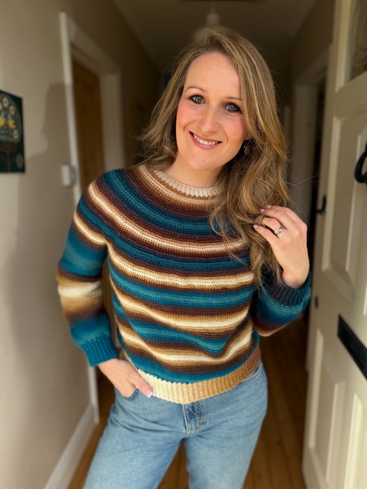 A woman standing indoors, smiling at the camera, wearing a striped crochet sweater in blue, brown, and beige tones with blue jeans.