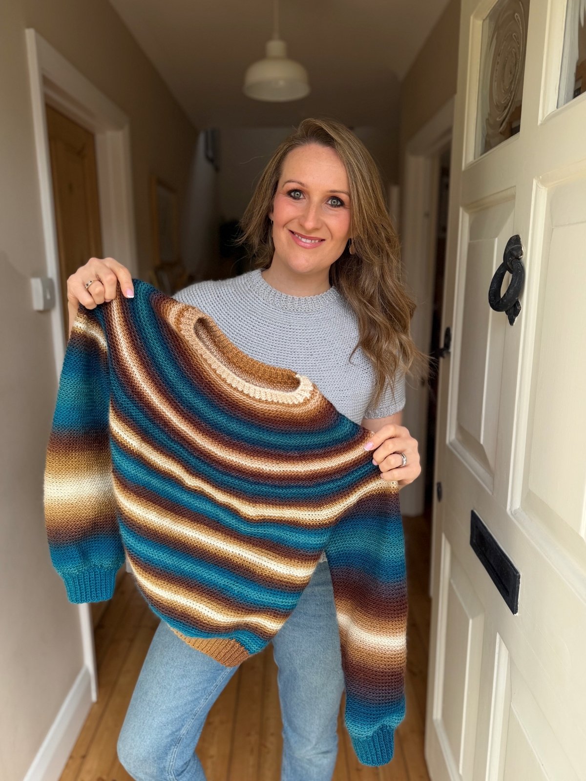 A woman stands in a hallway holding up a striped, multicolored crochet sweater, smiling at the camera.
