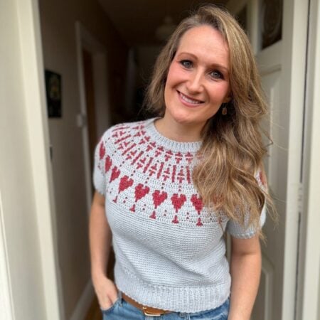 Woman standing in a doorway, smiling, wearing a light gray knit sweater with a red and white heart pattern and blue jeans.
