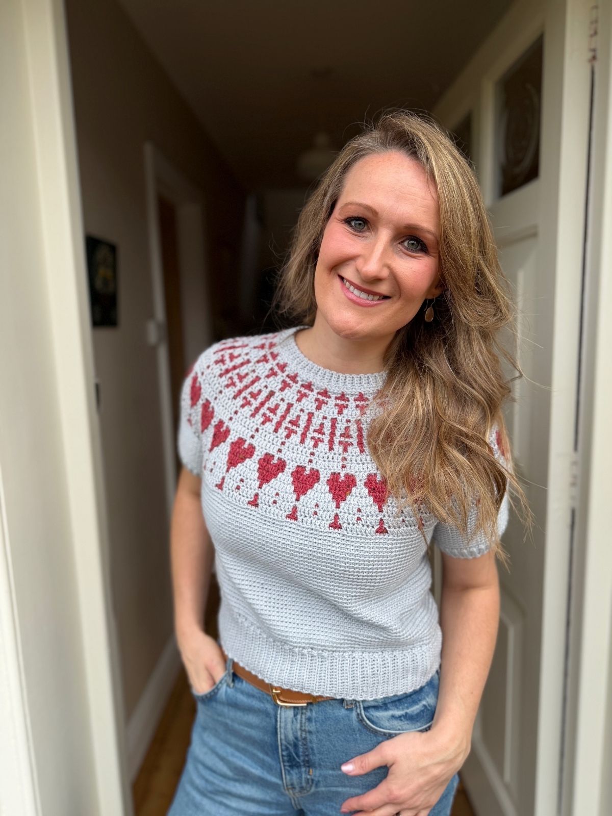 Woman standing in a doorway, smiling, wearing a light gray crochet sweater with a red and white heart pattern and blue jeans.