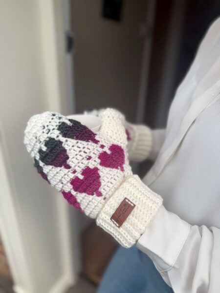 A person wearing white and multicolored crochet heart mittens holds a white cup. The cozy mittens feature a brown label on the wrist.