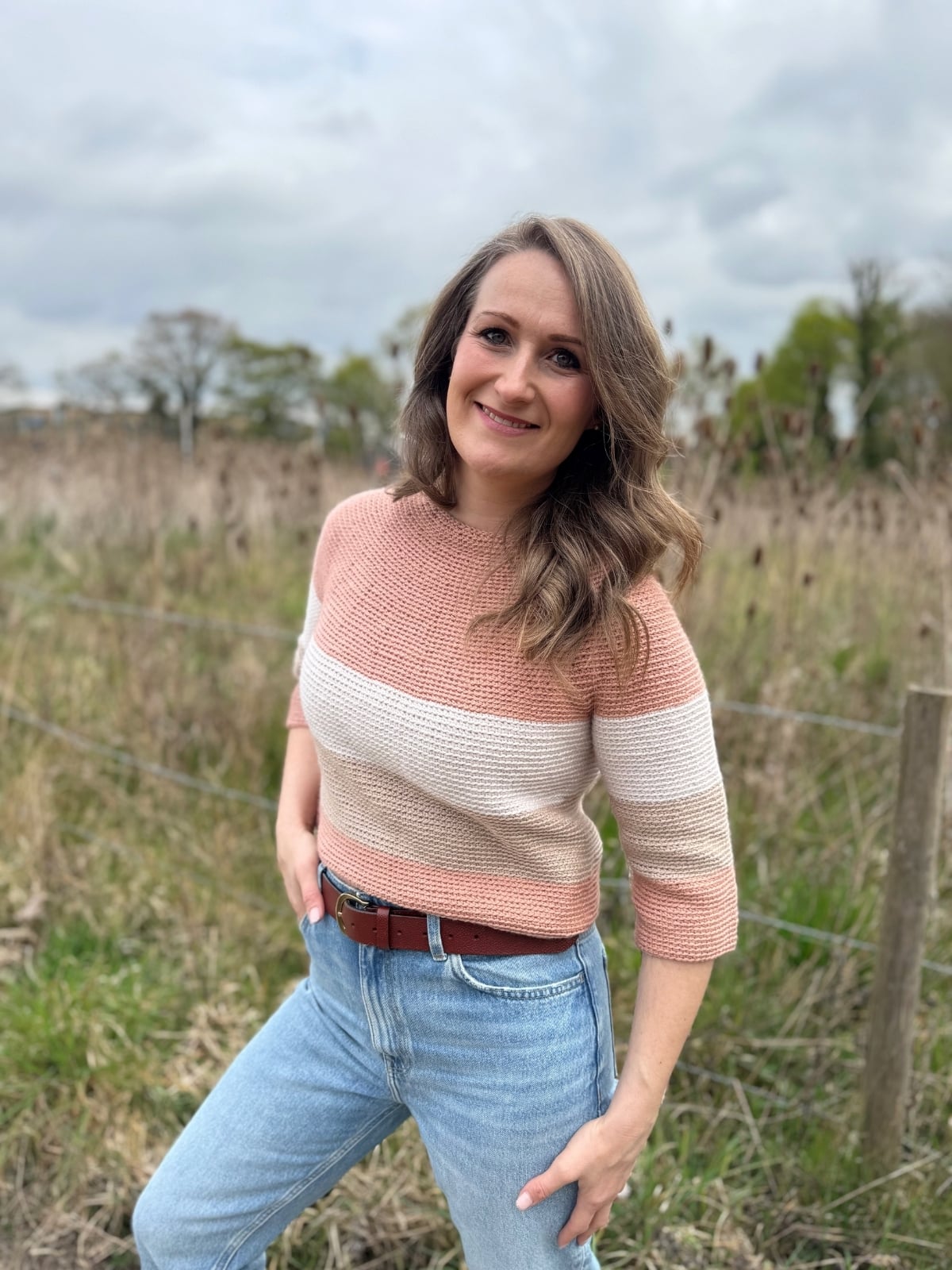 A woman with wavy hair stands outdoors in front of tall grass, wearing a striped crochet sweater and blue jeans, smiling at the camera.