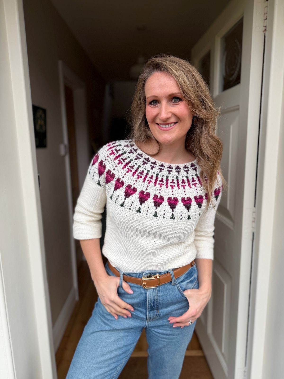 Woman standing in a doorway, wearing a white crochet sweater with a red and purple heart pattern and blue jeans, smiling at the camera.