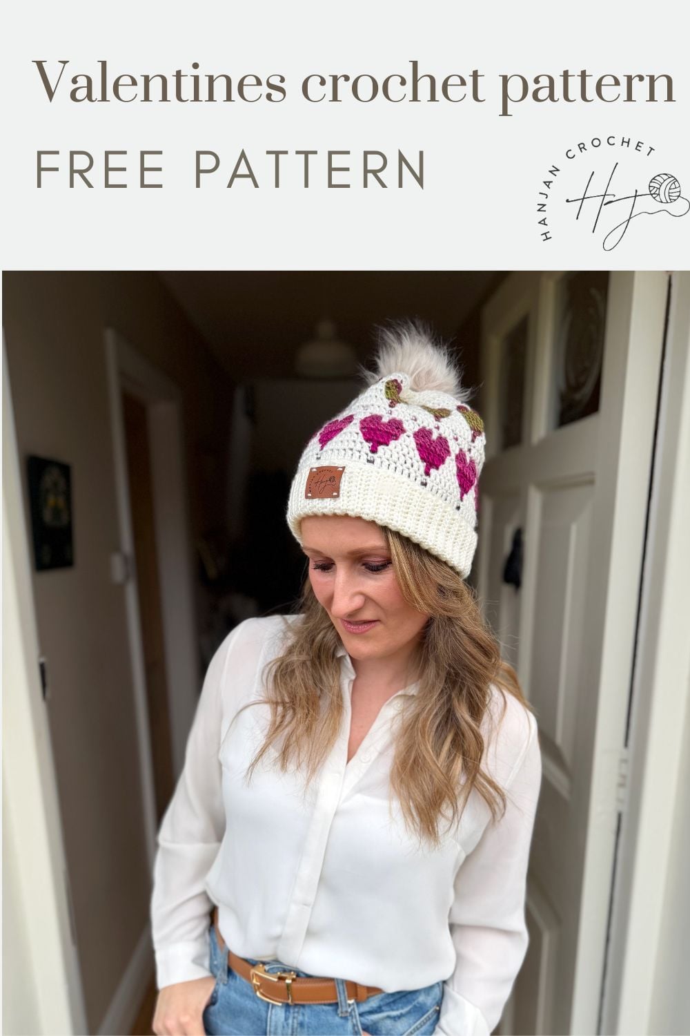 A woman in a white blouse and a crochet beanie with pink heart patterns stands indoors under a banner that reads "Valentines crochet pattern FREE PATTERN," showcasing the Free Crochet Heart Hat Pattern.