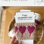 A hand holds a crochet coffee cozy with heart designs in pink, displayed on a "Happy Valentine's Day" insert. In the blurred background, a basket of yarn adds a cozy touch.