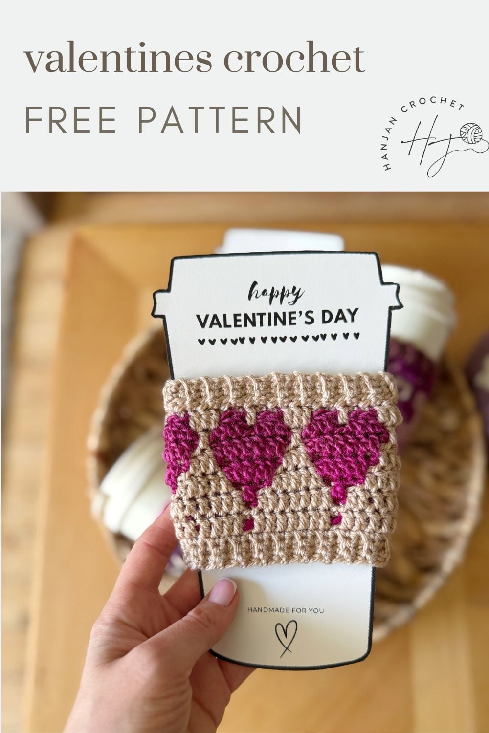 A hand holds a crochet coffee cozy with heart designs in pink, displayed on a "Happy Valentine's Day" insert. In the blurred background, a basket of yarn adds a cozy touch.