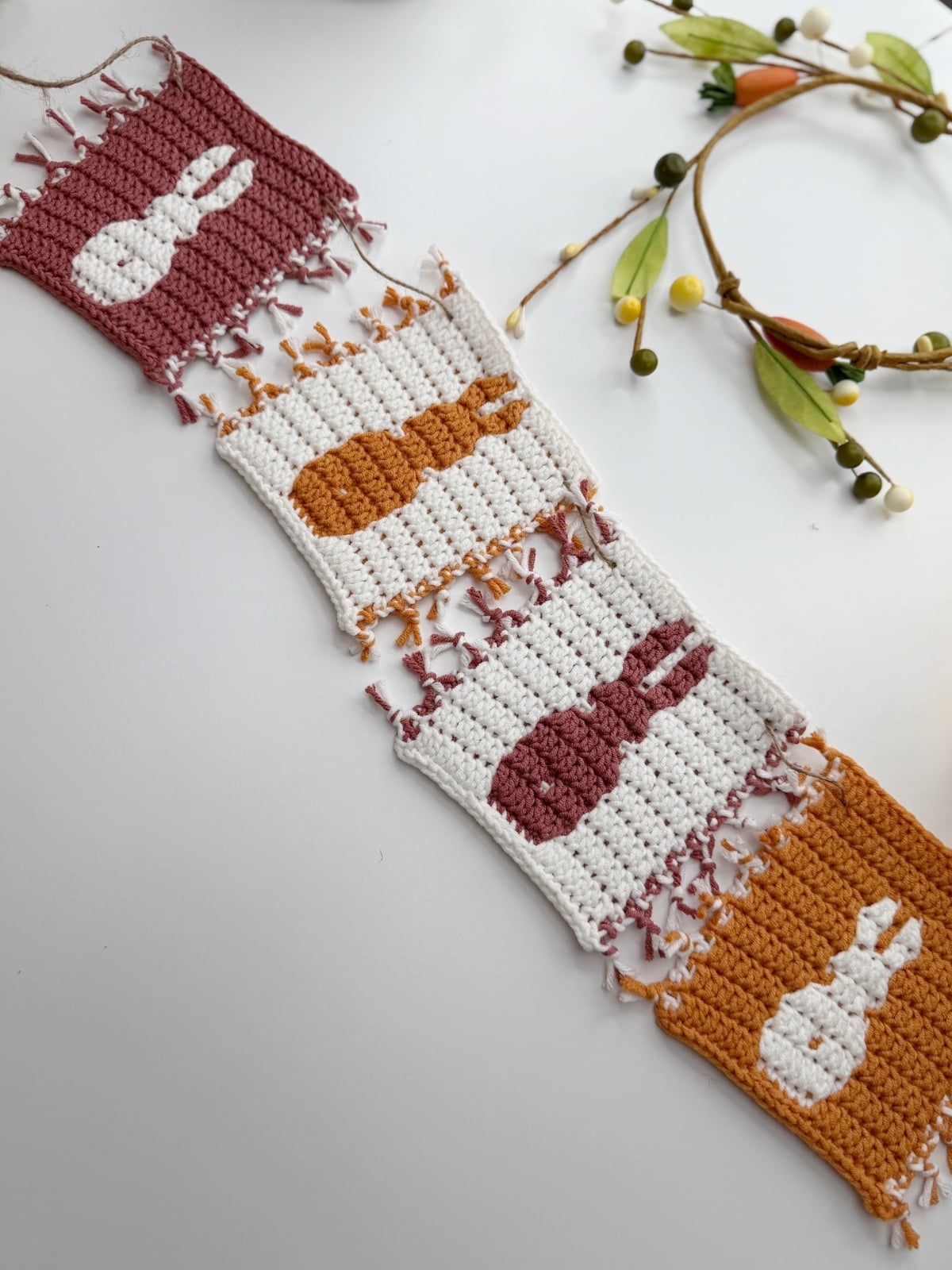 Four crocheted squares with bunny silhouettes in white, pink, and yellow yarn, laid out in a row on a white surface next to a decorative floral garland—perfect for showcasing your crochet bunny coaster pattern.