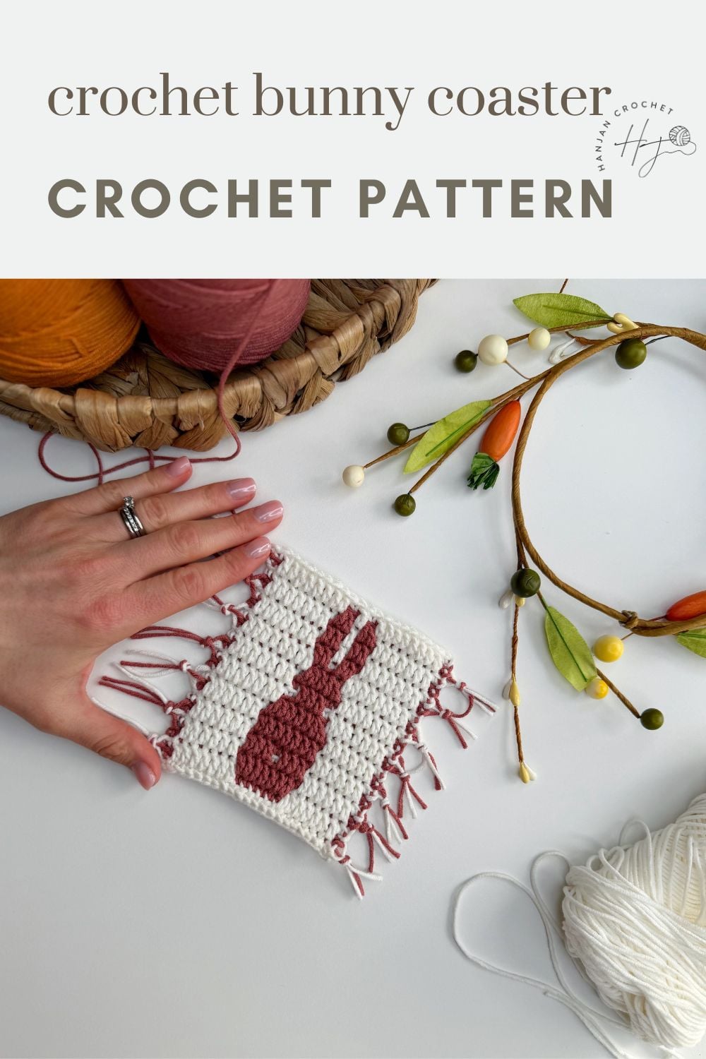 A hand holds a square crocheted coaster with a red bunny silhouette, surrounded by yarn, a basket, and a decorative branch. Text above reads, “crochet bunny coaster pattern CROCHET PATTERN.”.