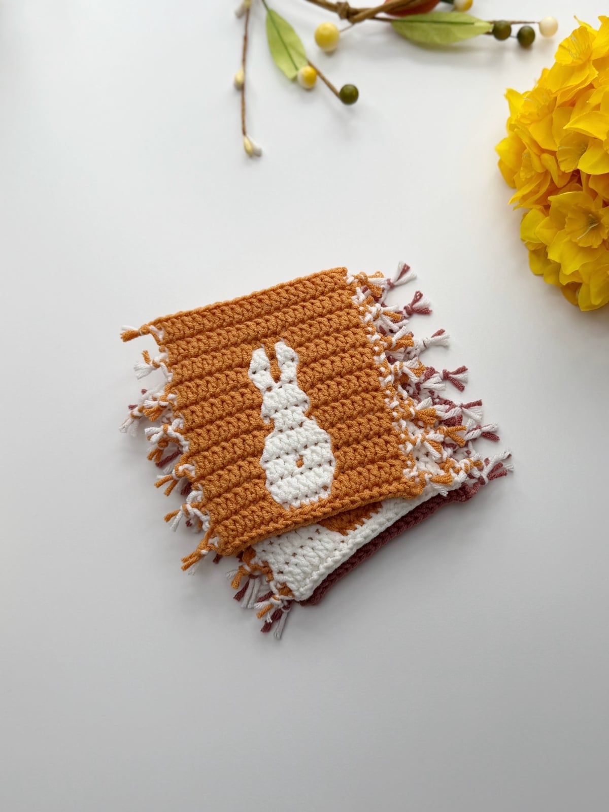 A crocheted square with a white bunny design and fringe edges, made using a crochet bunny coaster pattern, is placed on a white surface near yellow flowers and decorative stems.