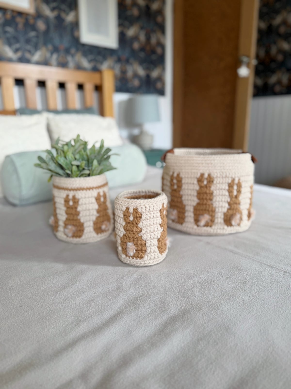 Three crocheted storage baskets, made with a charming crochet bunny basket pattern and one holding a small green plant, are arranged on a neatly made bed in a cozy bedroom.