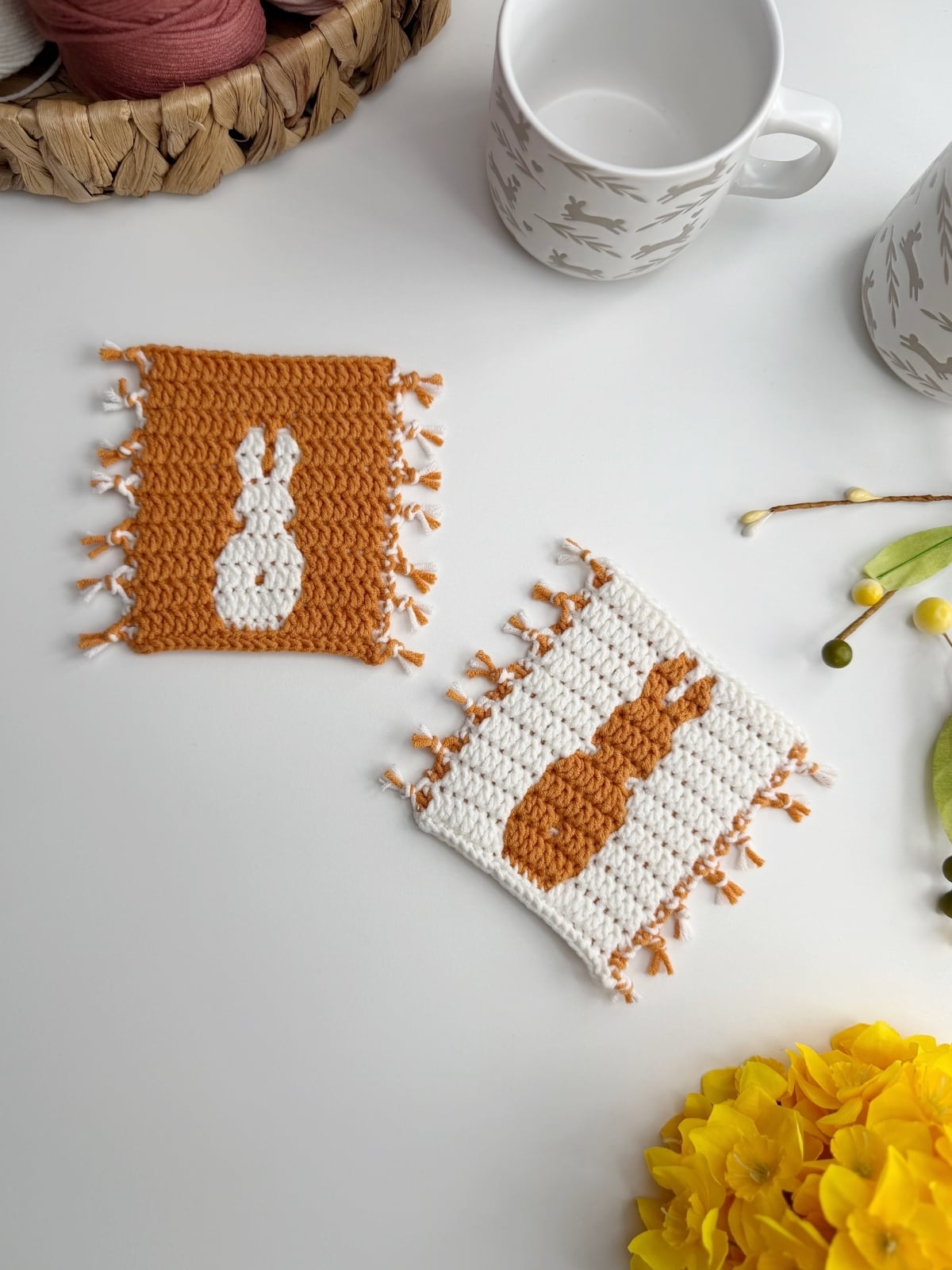 Two crocheted coasters with fringe, each featuring a bunny silhouette, are placed on a white surface beside cups, a basket with yarn, and yellow flowers.