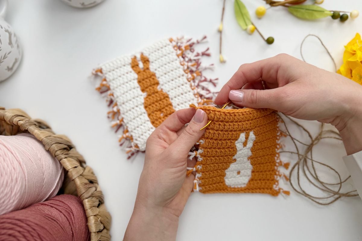 Hands crocheting a small orange square with a white bunny pattern; a finished Easter bunny crochet garland piece with an orange bunny on white is nearby, along with yarn and spring decorations.