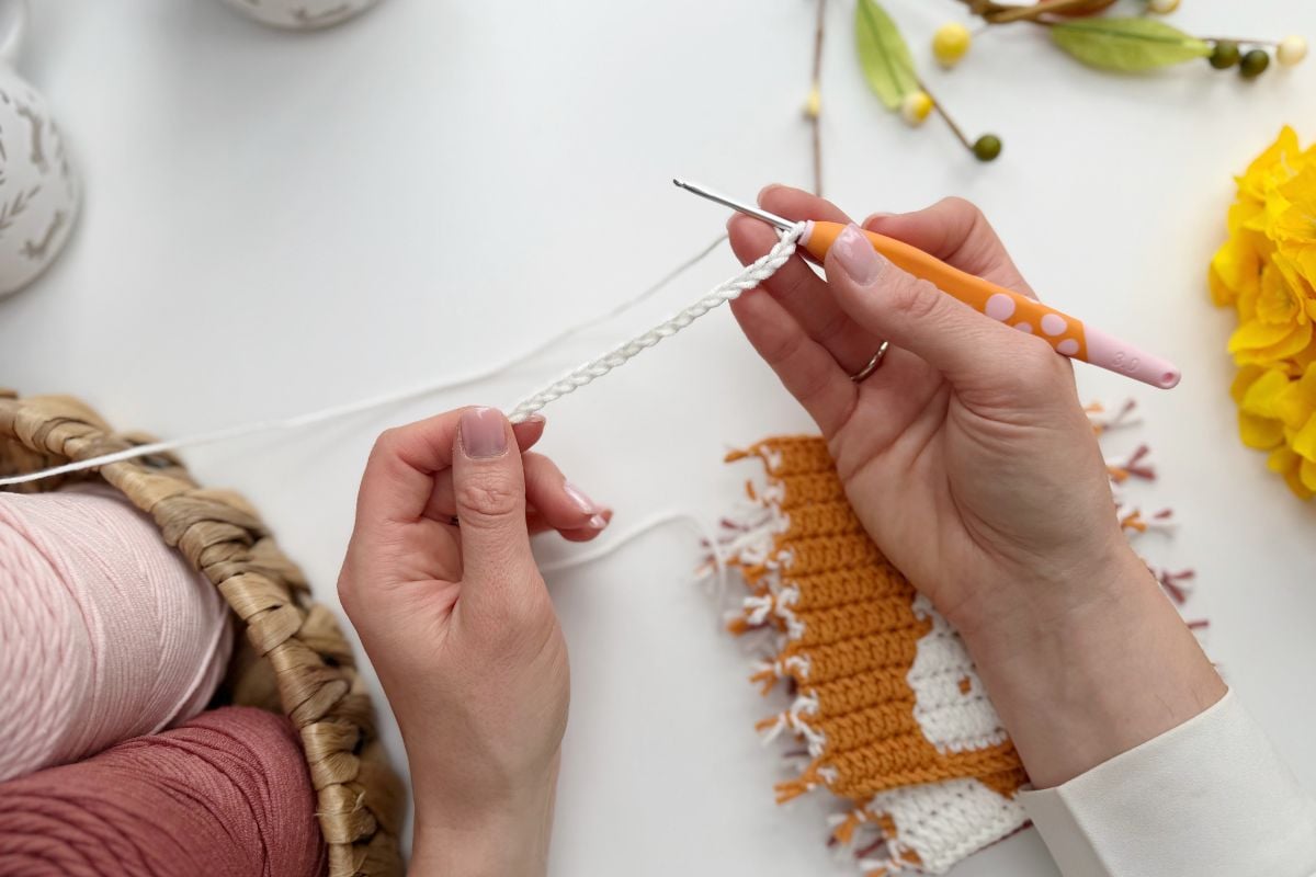 A person crochets with white yarn and an orange hook, creating an Easter bunny crochet garland, surrounded by yarn balls, a crochet piece, and decorative items on a white table.
