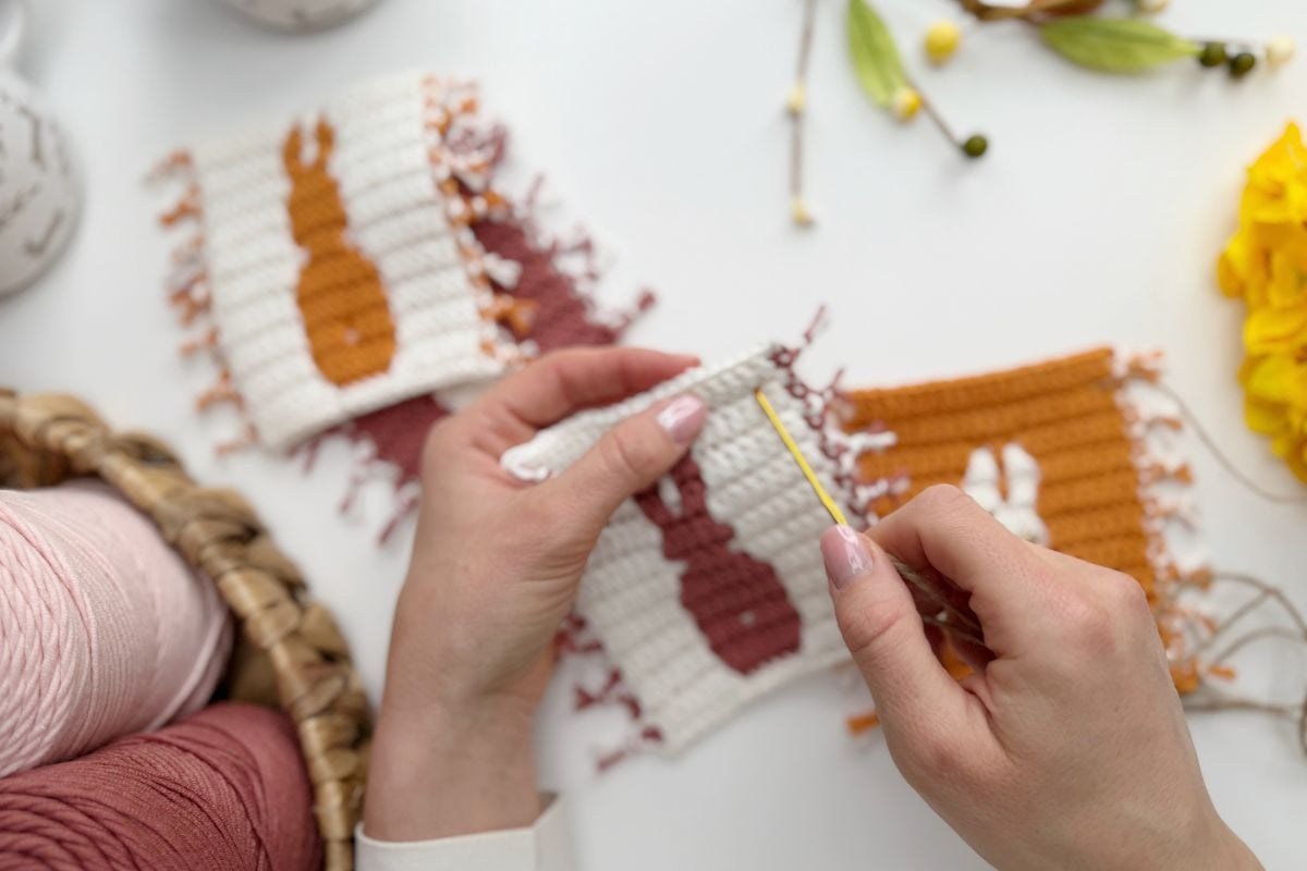 Two hands crochet a small square featuring an Easter bunny silhouette, with finished squares for a charming Easter bunny crochet garland, along with yarn and flowers on a white table.