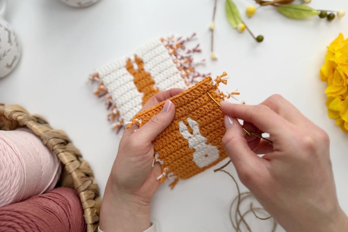 A person is hand-stitching a small crocheted piece featuring an Easter bunny silhouette on an orange background, with yarn and craft materials nearby&mdash;the perfect addition to a festive Easter bunny crochet garland.