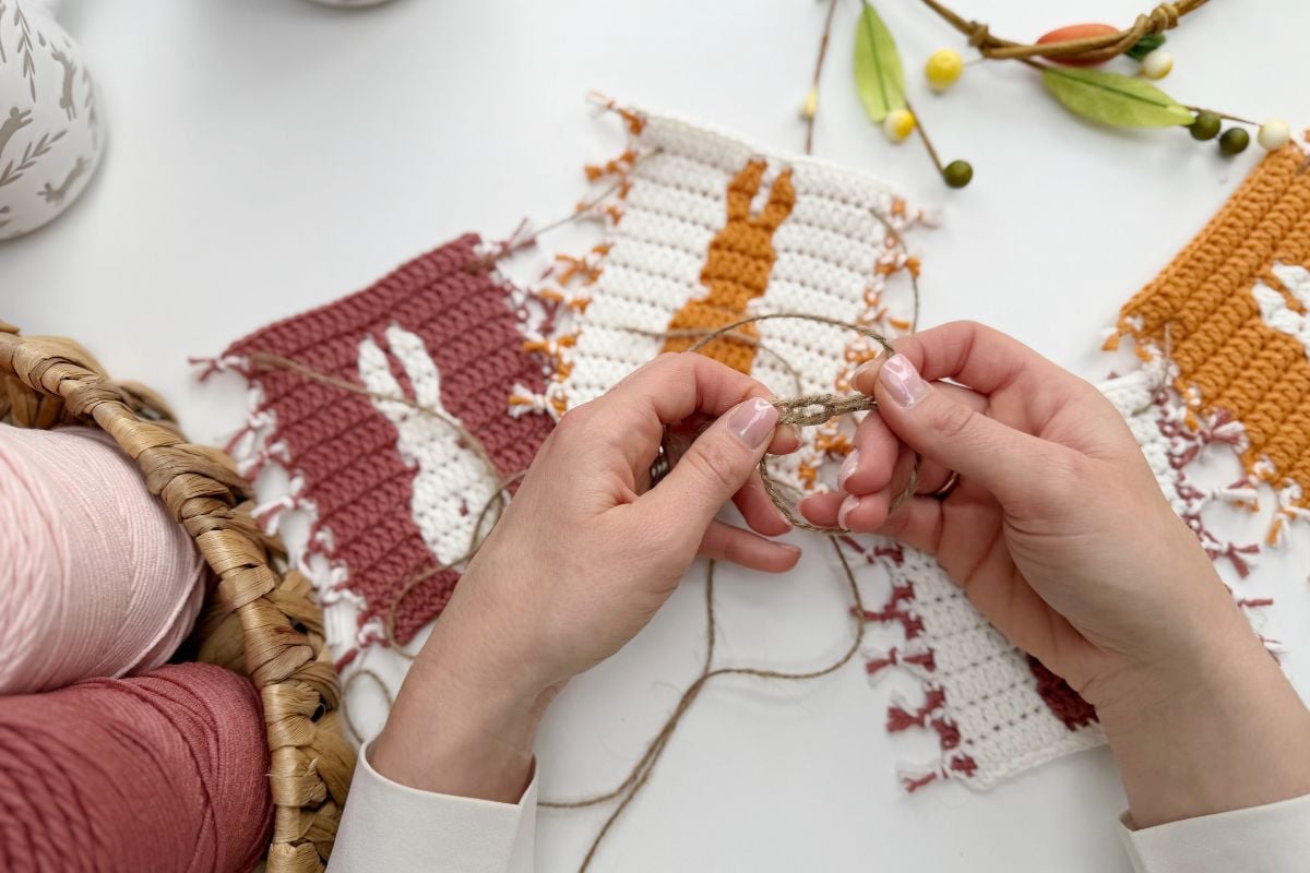 Hands crocheting a string with yarn, next to finished panels for an Easter bunny crochet garland in pink, white, and orange. A basket of yarn and decorative branches complete the festive scene.