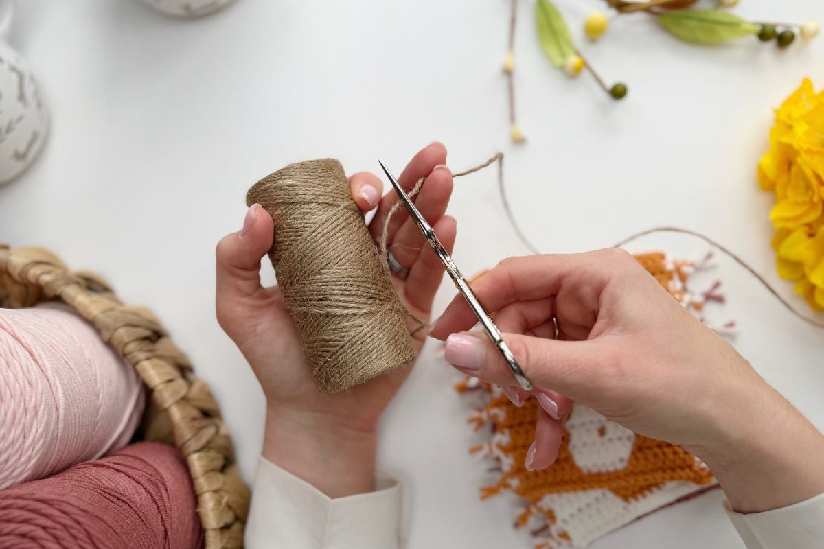 A person holds a spool of brown twine in one hand and cuts it with scissors, with yarn, crochet pieces, and an Easter bunny crochet garland alongside flowers on a white surface.