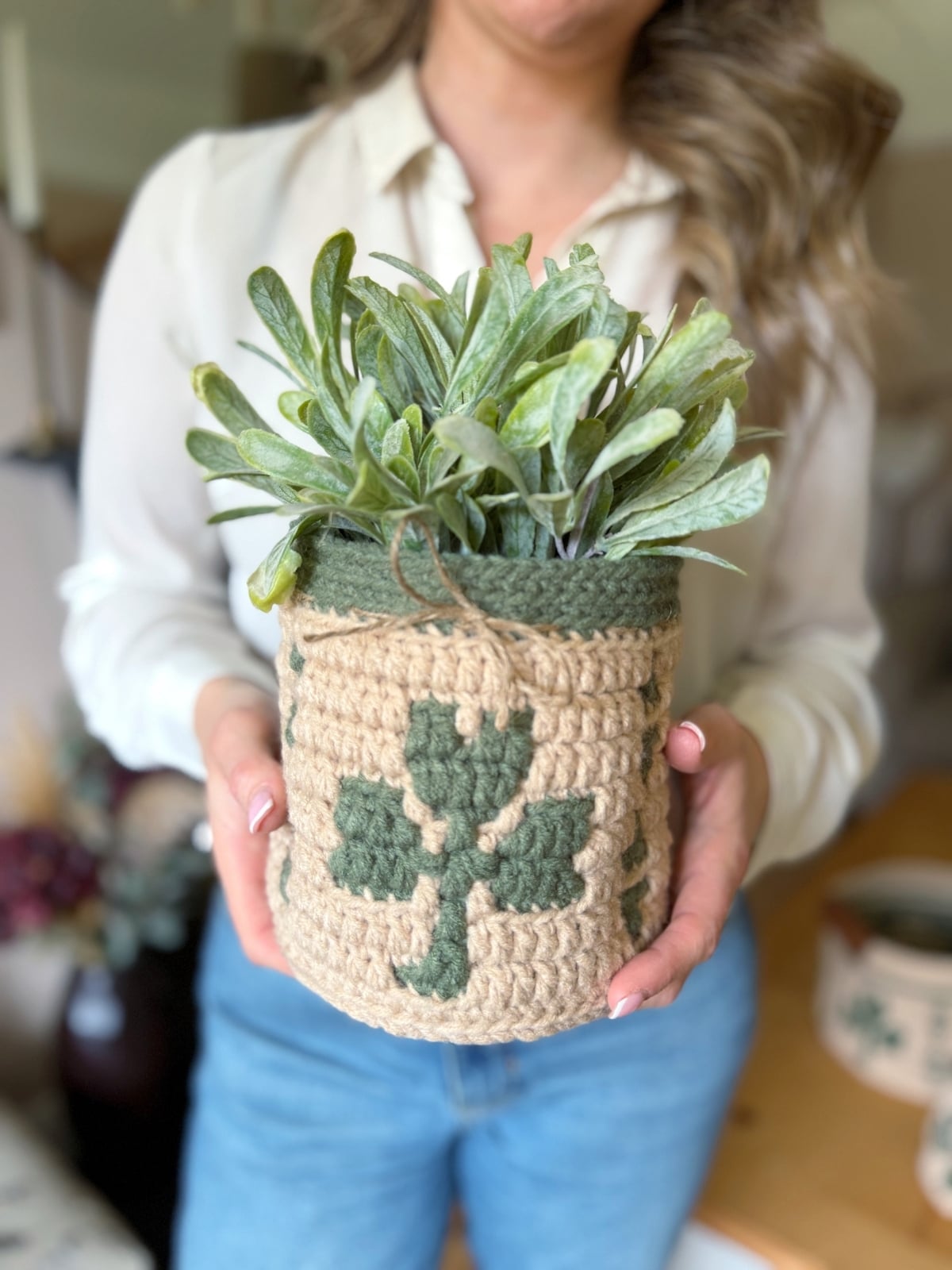 A person holding a potted plant in a beige and green crocheted planter featuring a charming crocheted shamrock design.