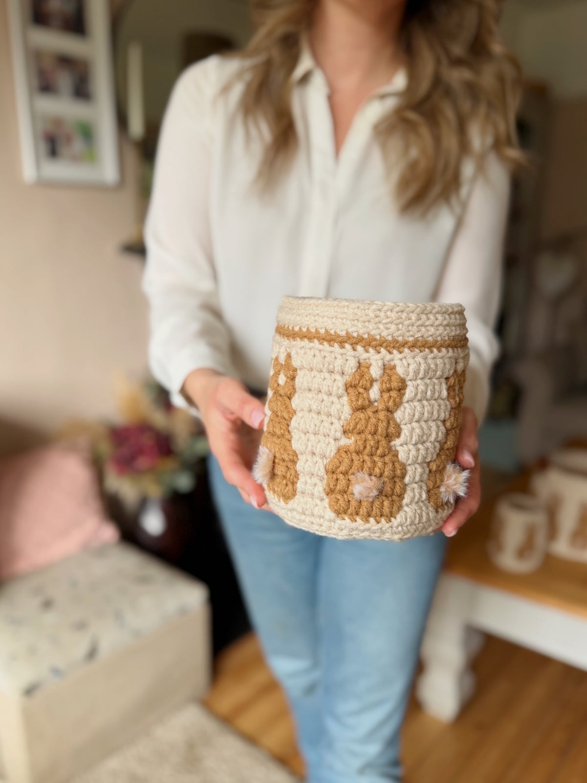 A woman in a white blouse and jeans holds a crocheted basket, made using a charming crochet bunny basket pattern, decorated with brown bunny silhouettes in a cozy living room.