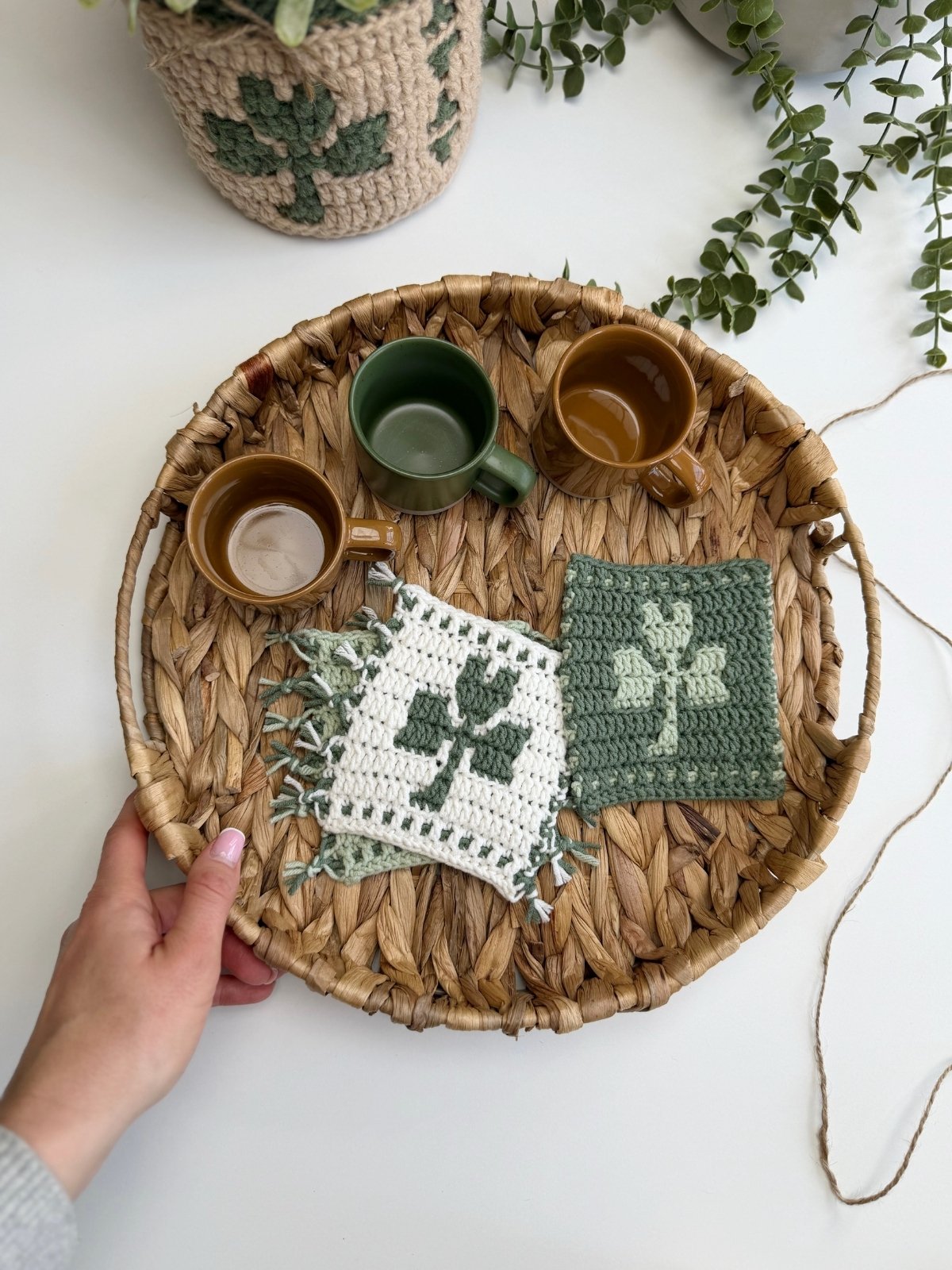A hand holds a woven tray with two brown cups, one green cup, and crocheted coasters featuring shamrock designs. Potted plants add a lush background to this cozy scene with a unique crochet shamrock coaster.