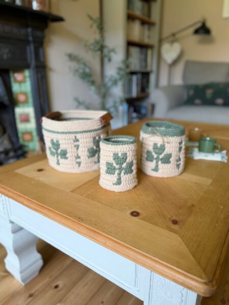Three crochet storage baskets with green leaf patterns, including a charming crocheted shamrock, are displayed on a wooden coffee table in a cozy living room setting.