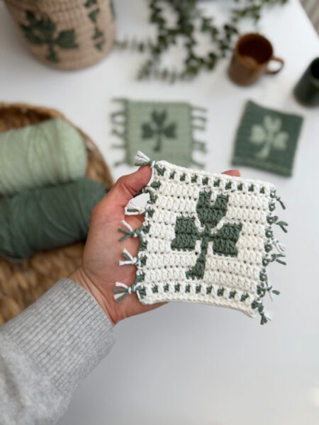 A hand holds a crochet shamrock coaster—a crocheted square with a green clover pattern and fringed edges; more yarn, finished squares, and mugs are visible in the background.