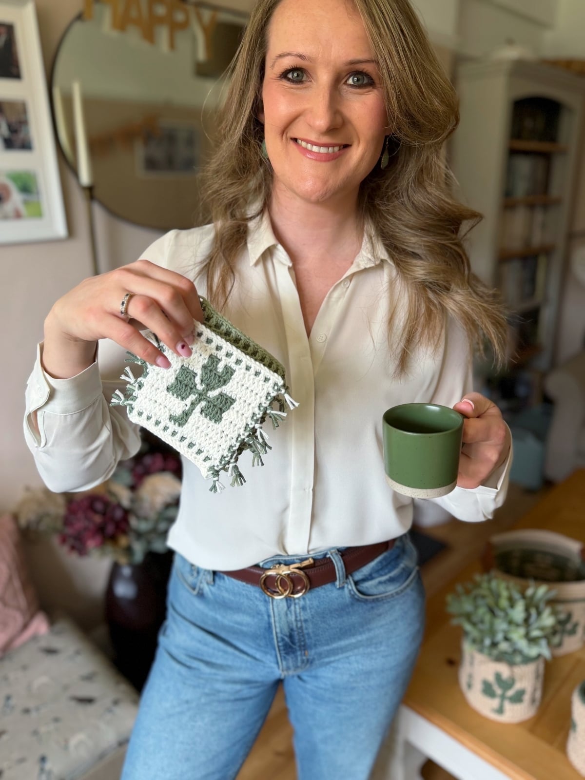 A woman in a white blouse and jeans holds a green cup and a crochet shamrock coaster with a shamrock design in a living room.