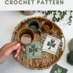 A hand holds a brown cup over a woven tray displaying three crochet shamrock coasters and another green cup; crochet yarn and plants are nearby.