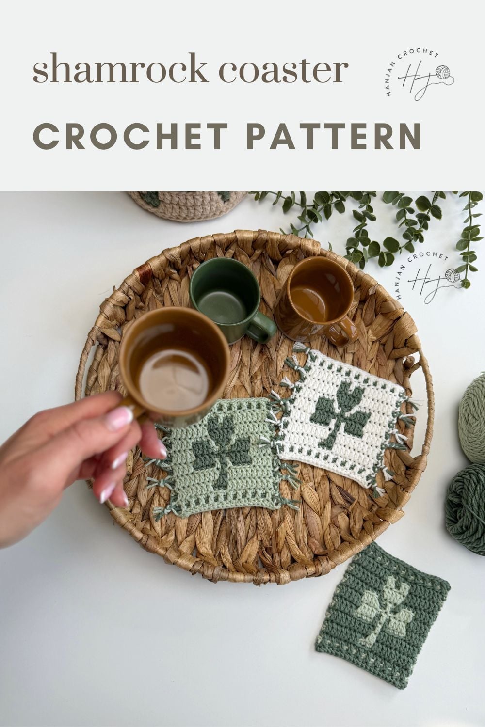 A hand holds a brown cup over a woven tray displaying three crochet shamrock coasters and another green cup; crochet yarn and plants are nearby.