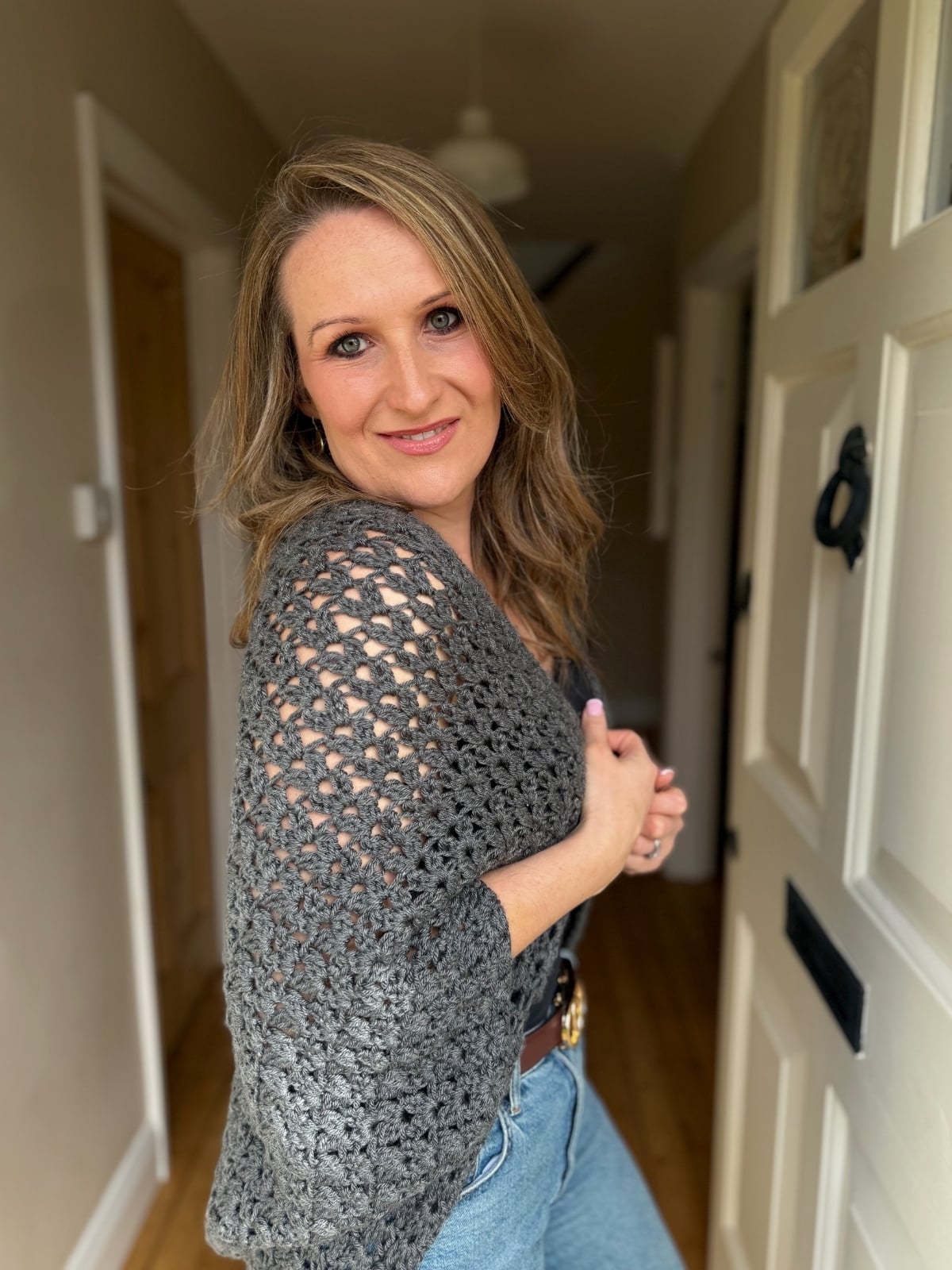 A woman with long, wavy hair stands in a hallway near an open door, wearing a gray crocheted shawl and blue jeans, smiling at the camera.