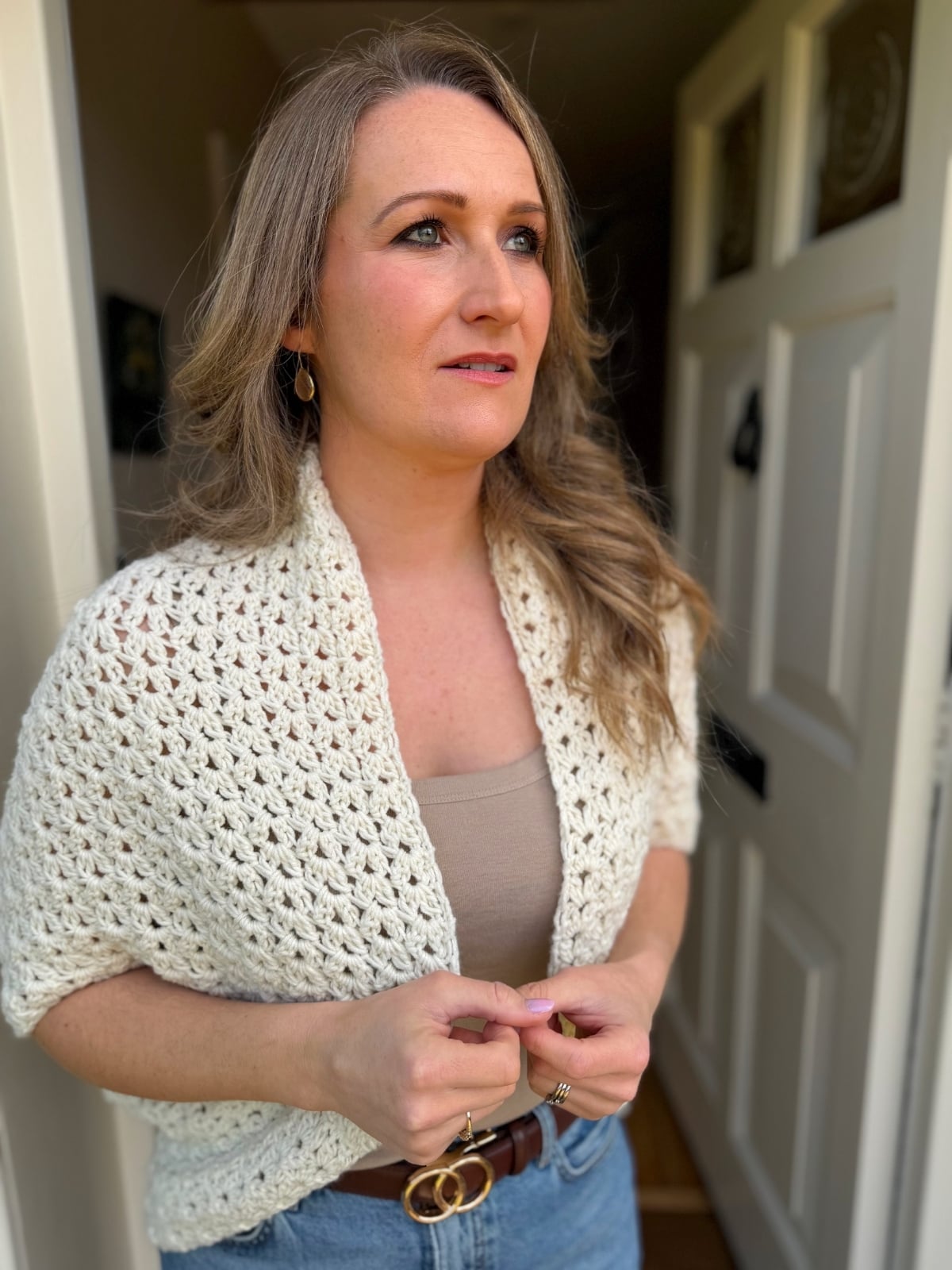 A woman with long, wavy blonde hair stands in a doorway, wearing a cream-colored crochet shawl, beige top, and blue jeans, looking slightly to the side.