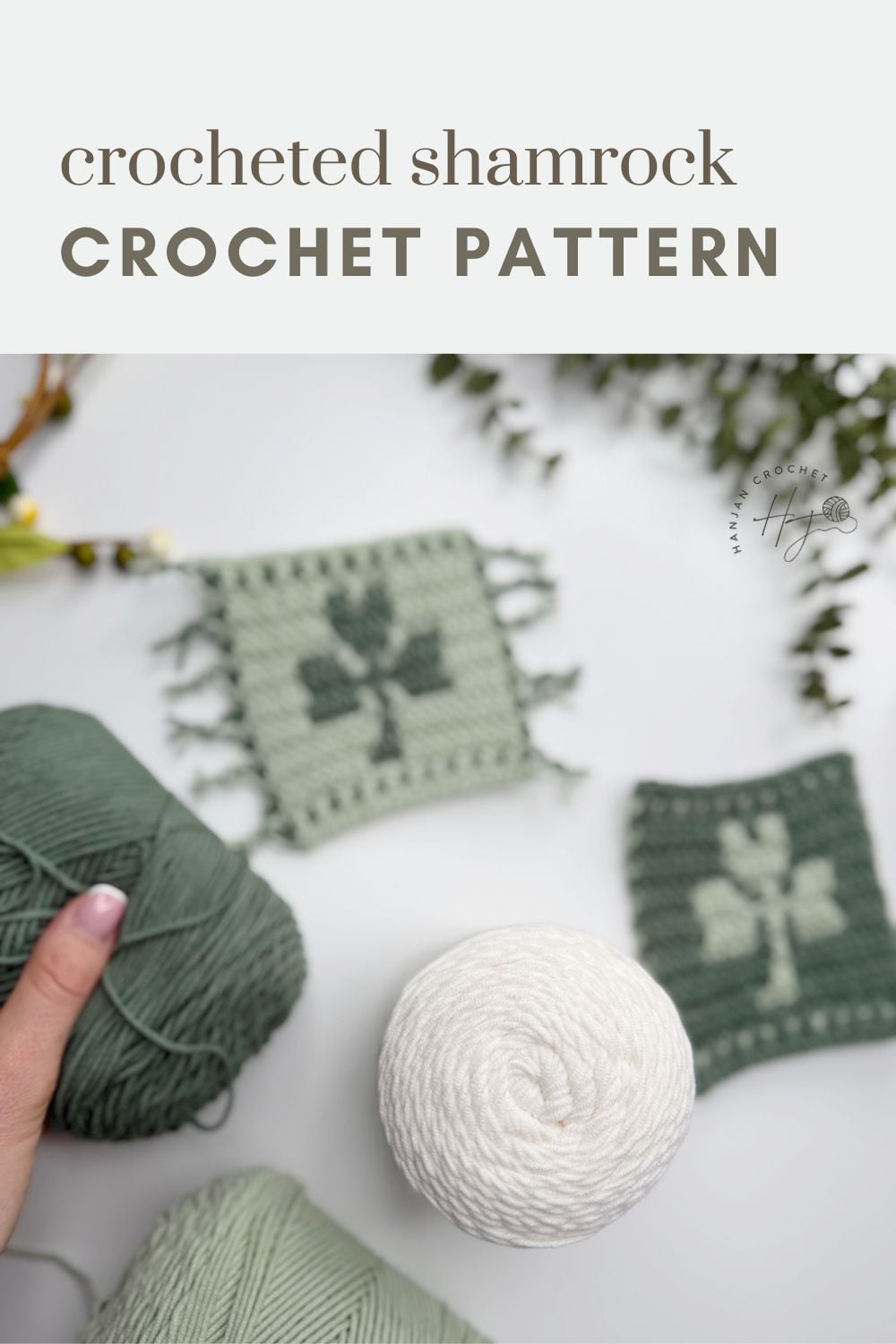 Three crochet shamrock coasters are displayed on a white surface, accompanied by green and white yarn balls and a hand holding green yarn.