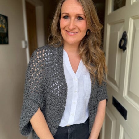 A woman with long blonde hair stands indoors by a white door, wearing a white blouse, grey knitted shawl, and black pants, smiling at the camera.