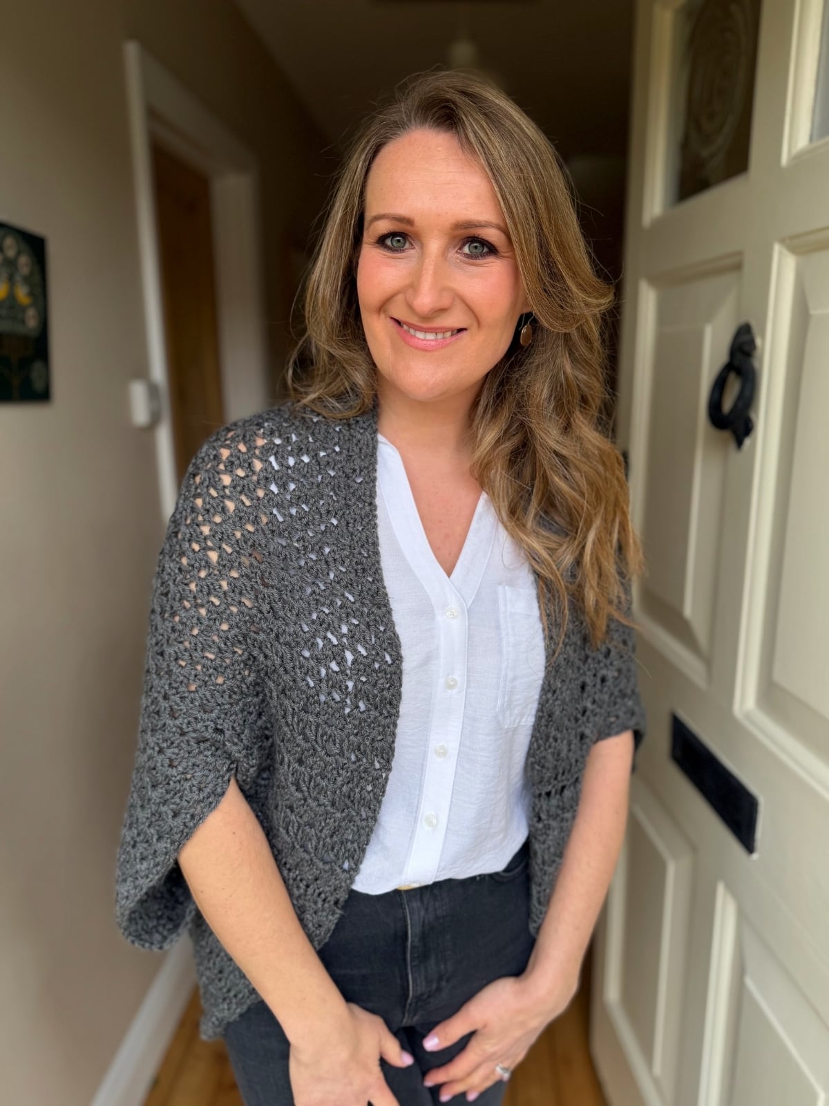 A woman with long blonde hair stands indoors by a white door, wearing a white blouse, grey knitted shawl, and black pants, smiling at the camera.