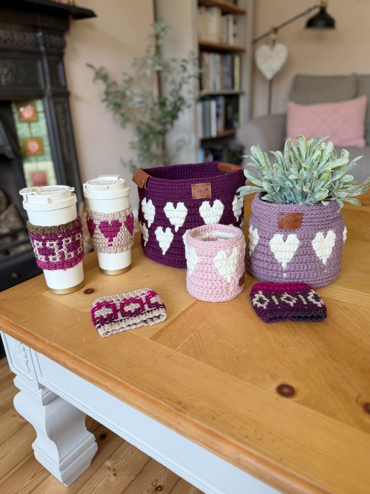 Various crocheted items with heart patterns, including a crochet heart basket pattern, cup sleeves, and a small pouch, are displayed on a wooden table in a cozy living room setting.