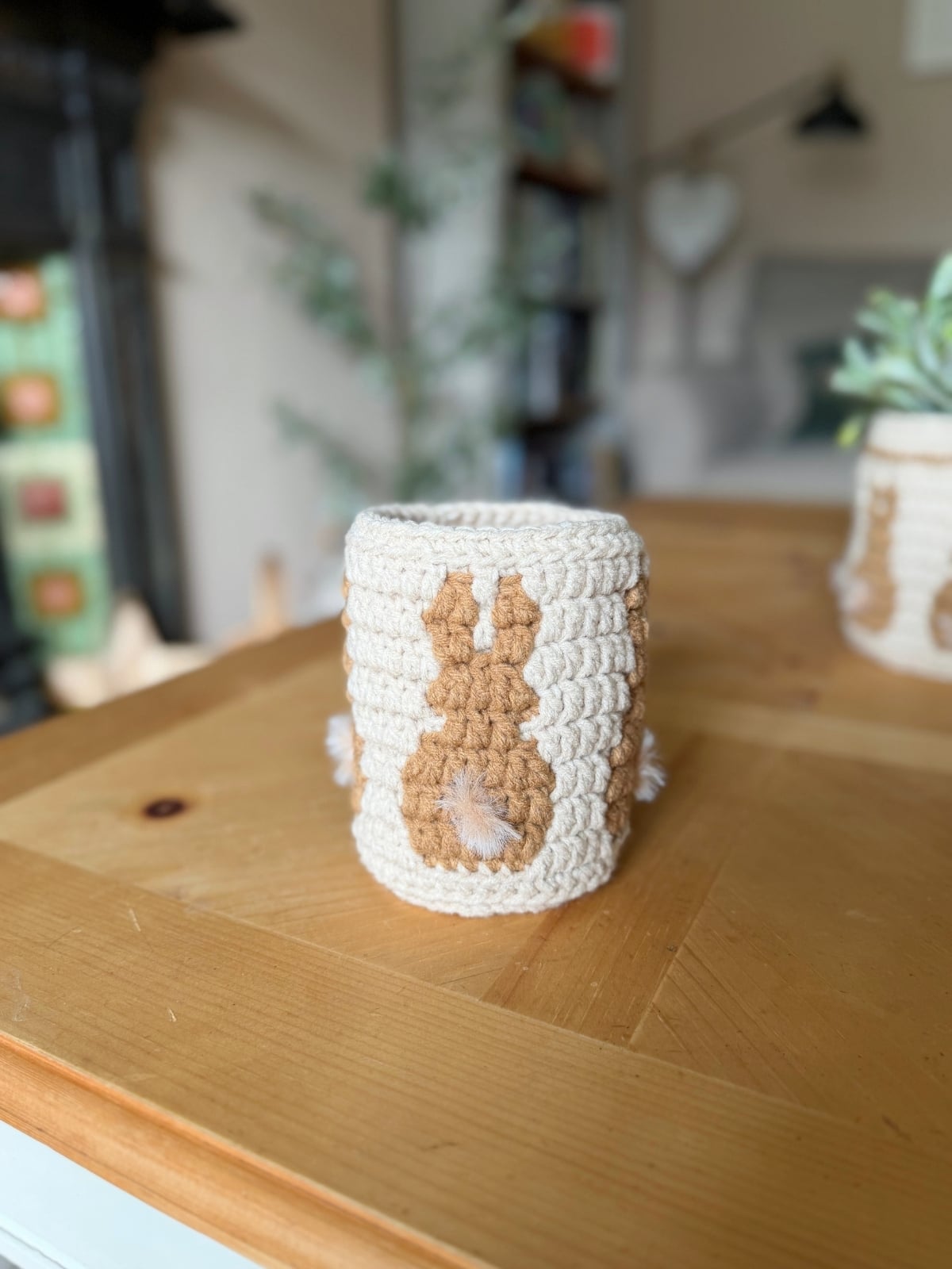 A crocheted beige and white cup cozy with a bunny design, inspired by a crochet bunny basket pattern, sits on a wooden table in a softly lit room.